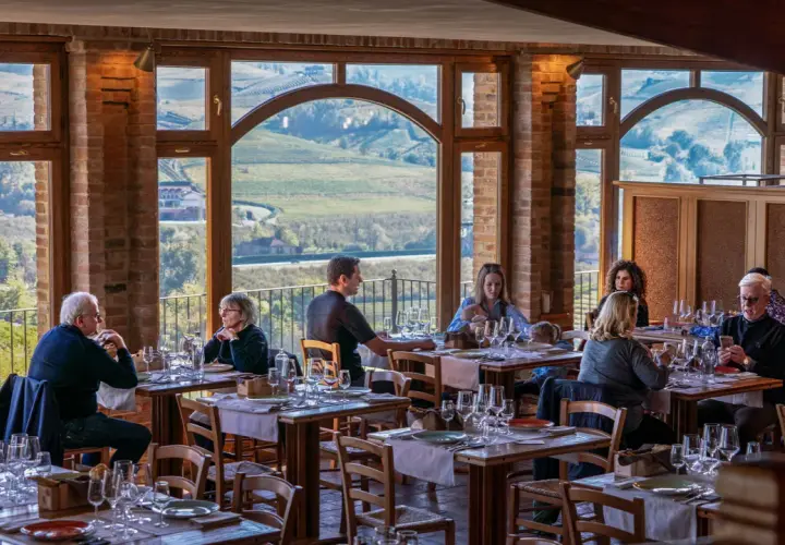 Persone cenano in un ristorante con vista panoramica su un'azienda agricola marrone. Tavoli apparecchiati con cura, mattoni a vista e grandi finestre creano un'atmosfera accogliente.