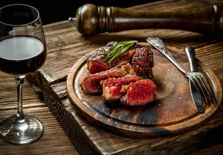 Bistecca succulenta con rosmarino, accompagnata da un bicchiere di vino tannico e posate d'argento su un tagliere di legno rustico.