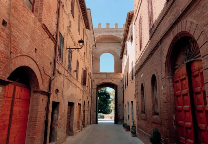 Un vicolo antico con edifici in mattoni rossi e un arco monumentale che conduce a un giardino. Sembra un buon convento.