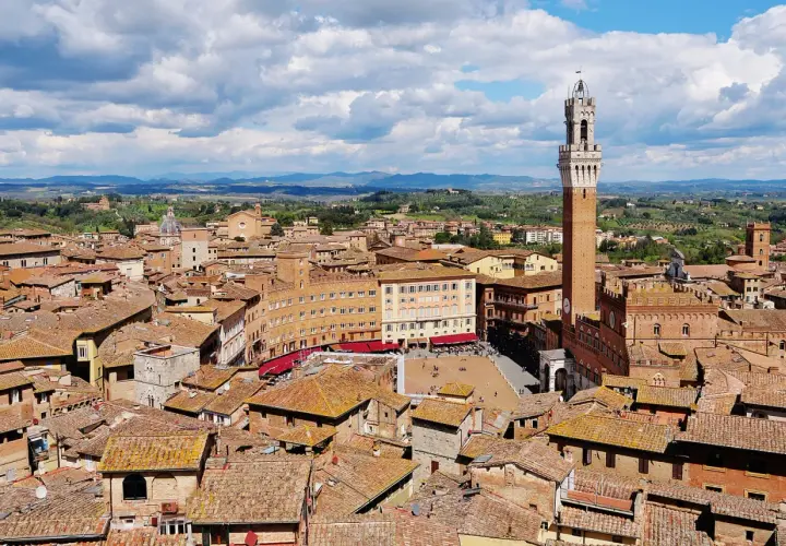 Vista aerea di Siena con la Torre del Mangia che domina la Piazza del Campo, circondata da tetti rossi e colline verdi.