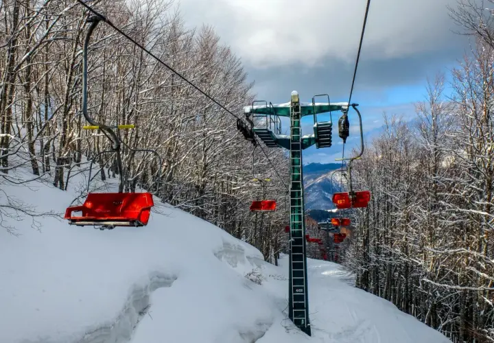 Seggiovie rosse e innevate salgono verso il cielo azzurro, attraversando un bosco spoglio. Un paesaggio invernale che ricorda il **centro** di **Abetone**.