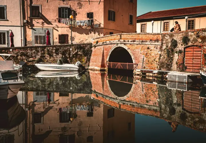 Un ponte in mattoni rossi attraversa un canale nel quartiere Venezia di Livorno. Barche ormeggiate riflettono sulle acque calme, mentre panni stesi aggiungono un tocco di vita quotidiana.