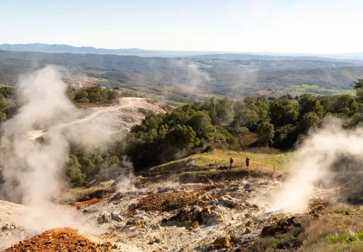 Vapori emergono dal terreno in un paesaggio collinare, con un villaggio in lontananza. Un geyser in Italia che incanta i visitatori.