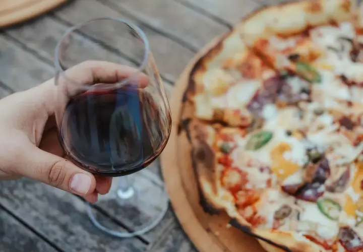 Un bicchiere di vino rosso in mano, accanto a una gustosa pizza. Un abbinamento perfetto per una serata all'aperto.