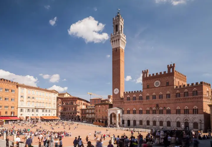 La Piazza del Campo di Siena, con la Torre del Mangia svettante, brulica di gente sotto un cielo azzurro.