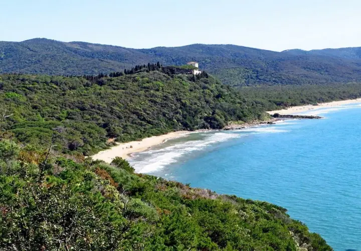 Spiaggia delle bandite di Scarlino, con mare azzurro, vegetazione lussureggiante e una villa in collina.