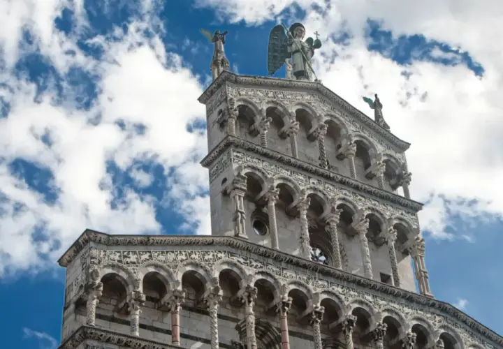 La facciata riccamente decorata della chiesa San Michele a Lucca, con un angelo in cima, si staglia contro un cielo azzurro e nuvoloso.