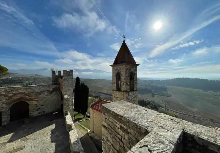 Torre campanaria e mura antiche del castello Frescobaldi, con vigneti e colline toscane sotto un cielo azzurro e soleggiato.