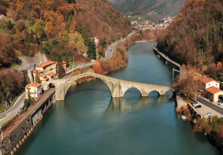 Il Ponte della Maddalena, con i suoi archi irregolari, attraversa un fiume circondato da colline autunnali e case colorate.