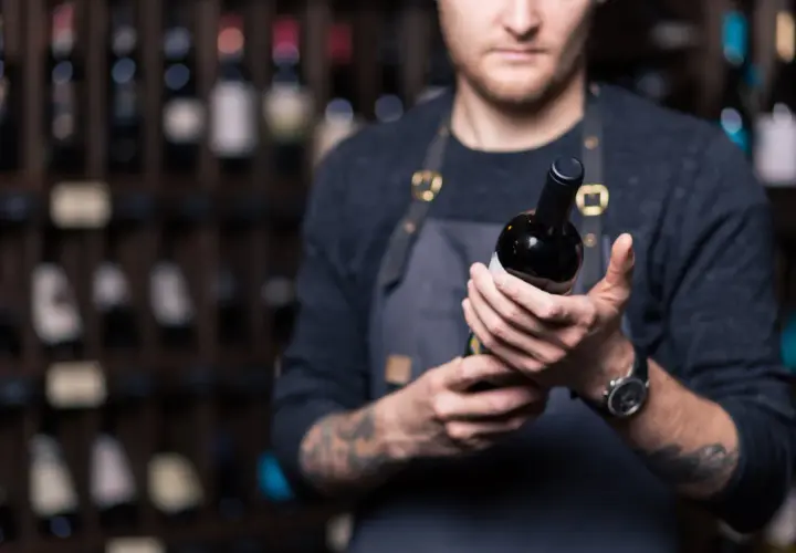 Un sommelier mostra una bottiglia di **vini toscani rossi pregiati**, con scaffali pieni di bottiglie sullo sfondo.