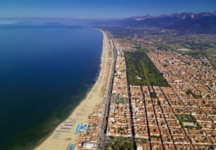 Vista aerea della costa di Massa Carrara, con il mare blu intenso, la lunga spiaggia attrezzata e la città che si estende verso le montagne.