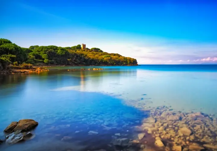 La costa verdeggiante con una torre antica, dove si trova Punta Ala, si specchia nelle acque calme e cristalline del mare.