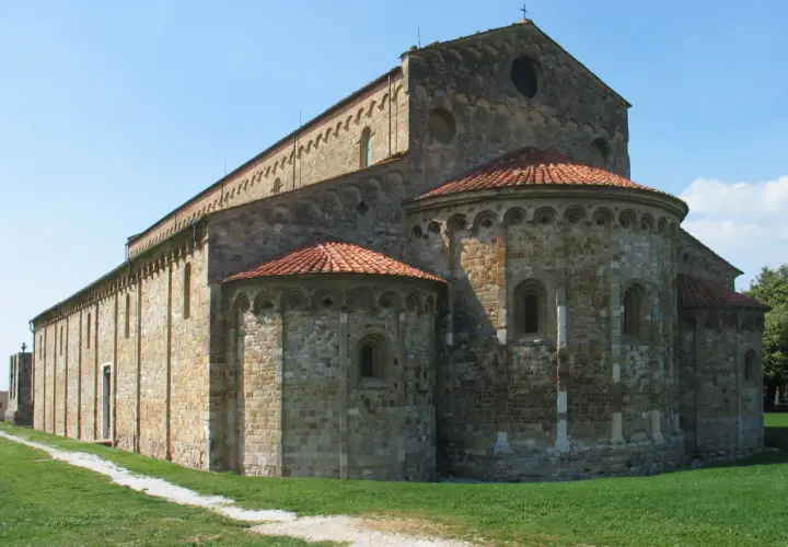 Basilica romanica di San Piero a Grado, con absidi semicircolari e tetto in tegole rosse, su un prato verde sotto un cielo azzurro.