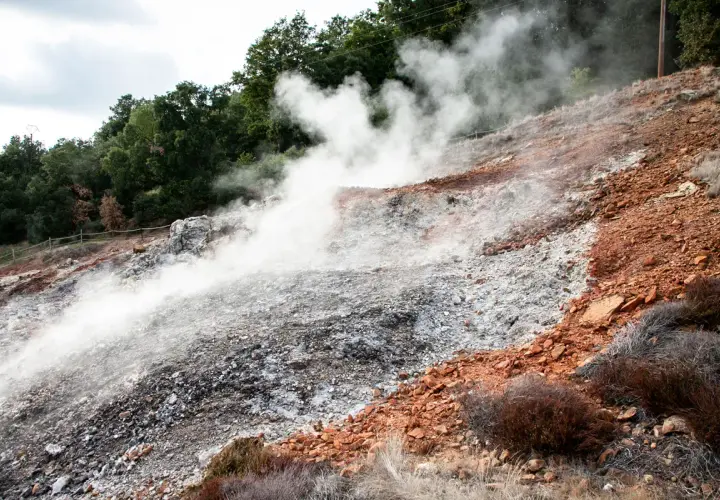 Fumarole di Monterotondo Marittimo emettono vapore su un pendio roccioso e arido, con vegetazione secca e alberi sullo sfondo.