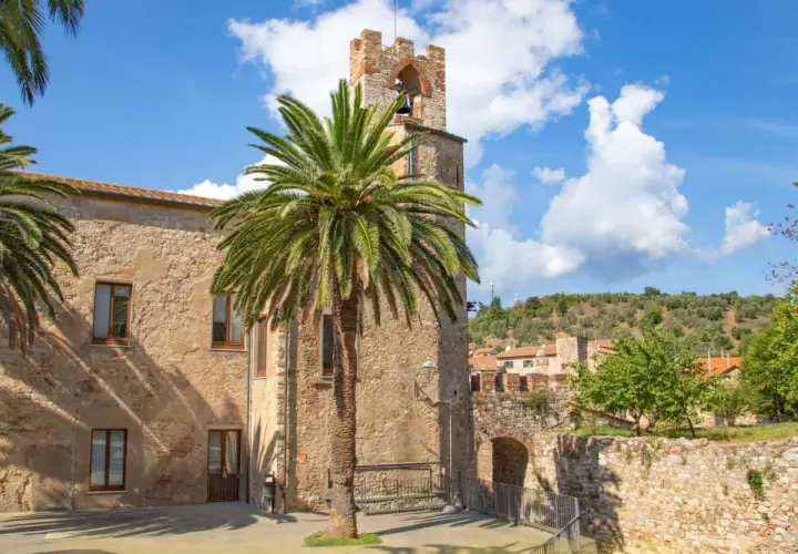 Un antico campanile in pietra a Suvereto, Livorno, svetta tra palme rigogliose e un cielo azzurro con nuvole bianche.