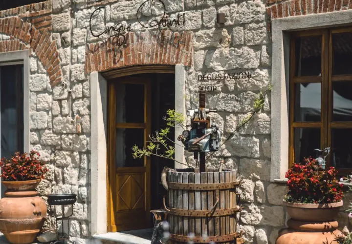 Ingresso di "Cinque Quinti", un negozio di degustazione con un torchio antico e vasi di fiori rossi.