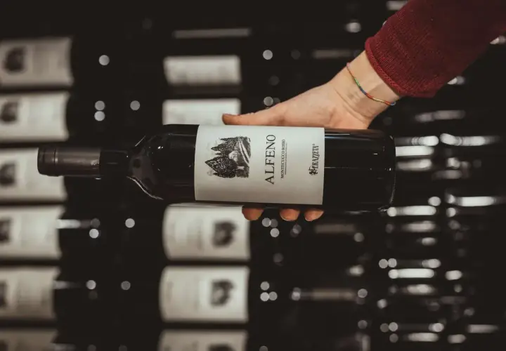 Mano che tiene una bottiglia di vino rosso, etichetta "Alferno Montecucco Rosso", un vino che ricorda il cabernet.