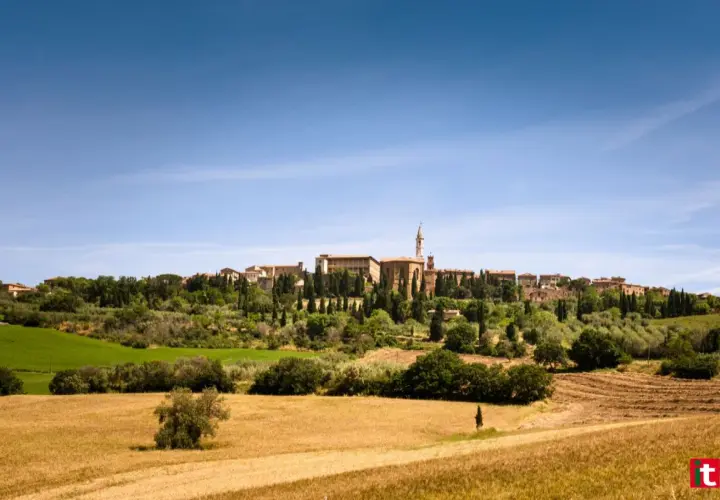 Un borgo medievale domina la Val d'Orcia, circondato da campi dorati e cipressi sotto un cielo azzurro.