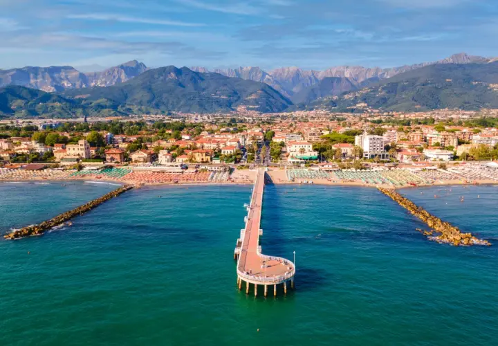 Un molo si estende nel mare turchese di Marina di Massa, con spiagge affollate e montagne sullo sfondo.