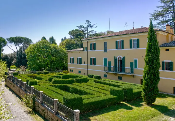 Villa Gracciano della Seta: un giallo edificio con un giardino geometrico di siepi verdi e un cipresso slanciato.
