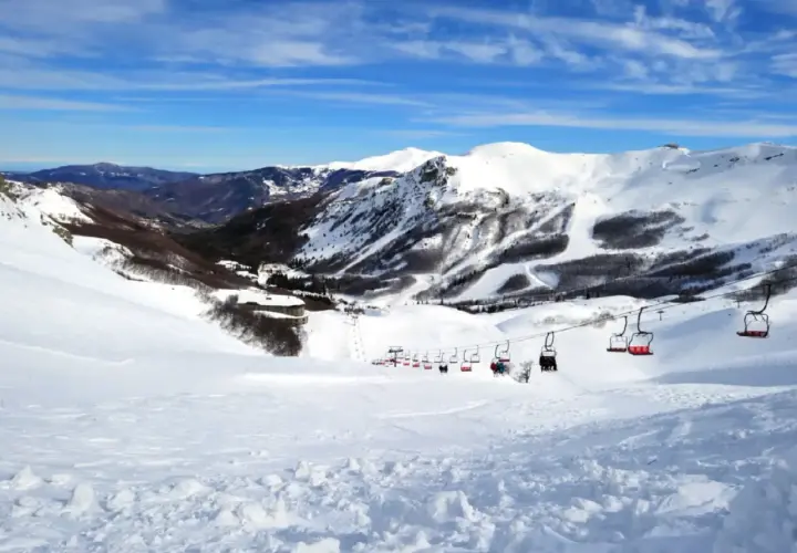 Impianti di risalita rossi trasportano sciatori su piste innevate ad Abetone, con montagne sullo sfondo sotto un cielo azzurro.