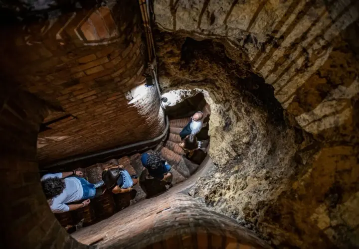 Persone esplorano le antiche cantine storiche di Montepulciano, scendendo una scala a chiocciola in mattoni e tufo.
