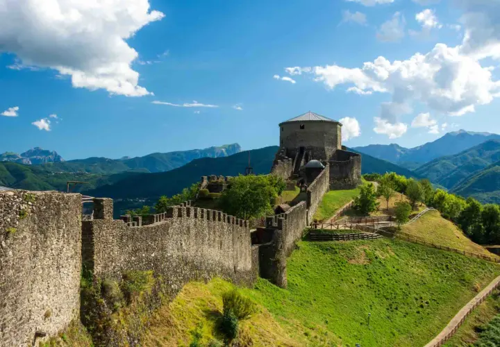 Roccia imponente e mura antiche del **San Romano in Garfagnana** si stagliano su un paesaggio verdeggiante, sotto un cielo azzurro con nuvole bianche.