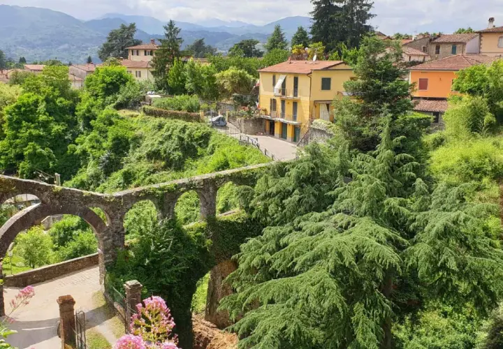 Un antico acquedotto in pietra, ricoperto di vegetazione, domina il paesaggio collinare dove si trova Barga. Case gialle e arancioni si stagliano tra il verde lussureggiante.