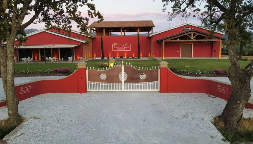 L'ingresso della Tenuta di Vaira a Bolgheri, con cancelli in rame e bianco, edifici rossi e un prato curato.