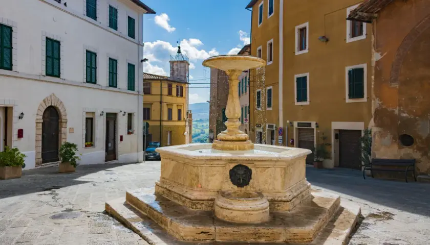 Fontana in pietra nel cuore dell'entroterra toscano, con palazzi storici e scorcio collinare.