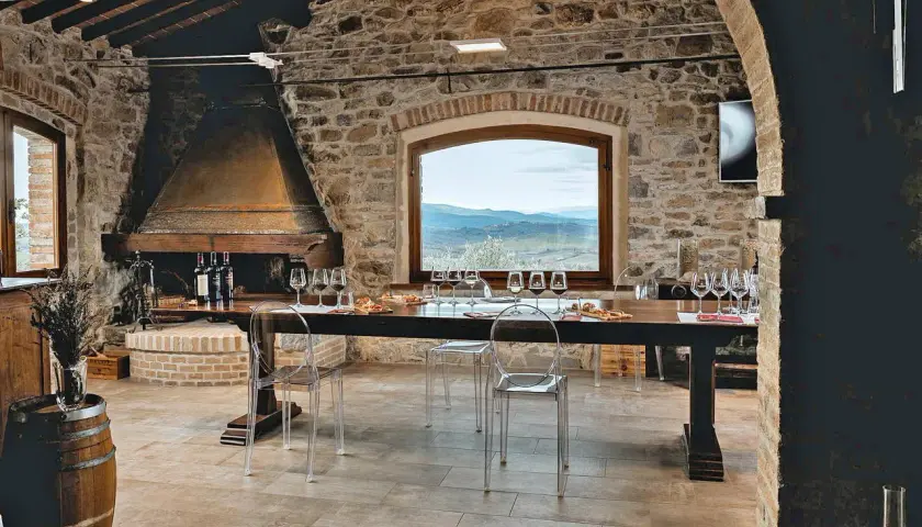 Sala degustazione in pietra con camino, tavola apparecchiata per assaggio vini, vista panoramica sulle colline del Chianti.