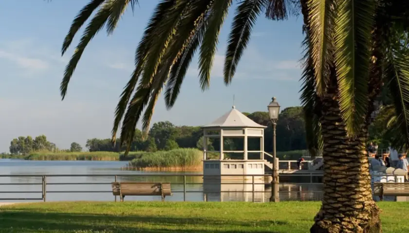 Un gazebo bianco si affaccia sul lago, vicino a una torre. Palme e panchine completano la scena del centro storico.