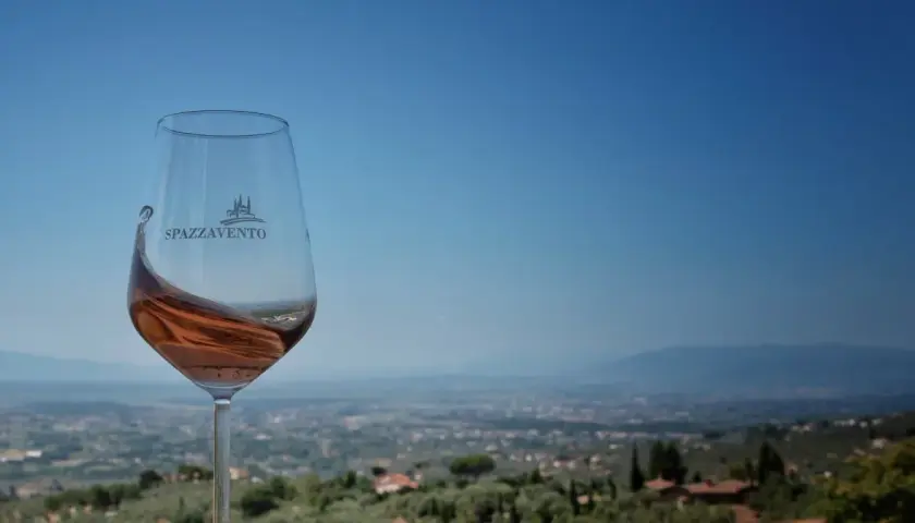 Calice di vino rosato con logo Spazzavento, sullo sfondo il panorama della fattoria di Piazzano e le colline toscane.
