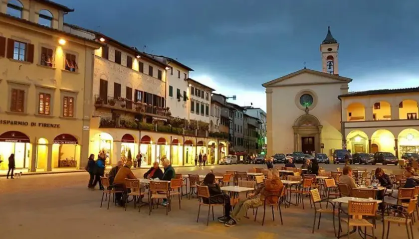 Serata in piazza a Figline Valdarno, con tavolini all'aperto e la chiesa illuminata.