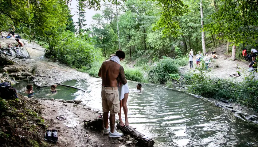 Persone si godono le acque termali ai Bagni San Filippo, un'oasi naturale in un bosco rigoglioso. Nonostante il nome, l'immagine evoca un'atmosfera estiva, non invernale.