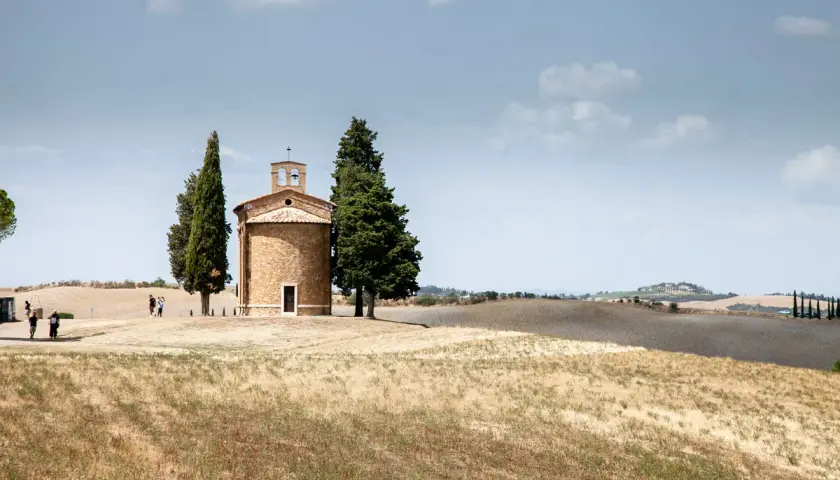 Iconica chiesetta Val d'Orcia, con cipressi e colline ondulate sotto un cielo nuvoloso.
