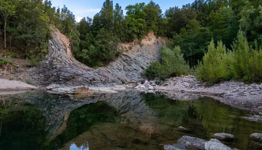 Rocce stratificate e vegetazione lussureggiante si specchiano nelle acque tranquille del Val di Farma, un paesaggio naturale mozzafiato.