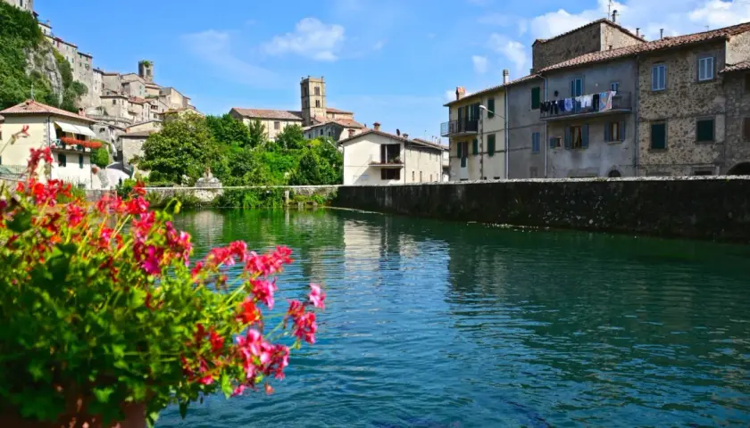 Fiori rossi in primo piano, con il borgo di Santa Fiora, Grosseto, che si specchia nell'acqua limpida.