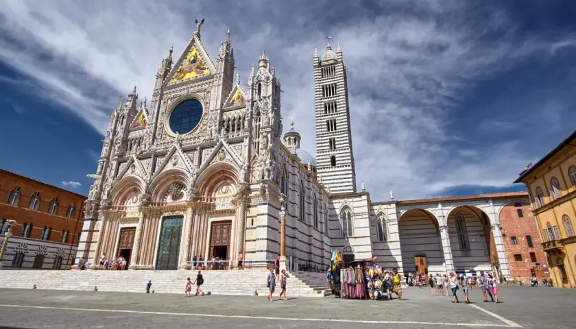 Magnifica facciata del Duomo di Siena, con la sua torre a strisce bianche e nere. Turisti passeggiano nella piazza antistante.