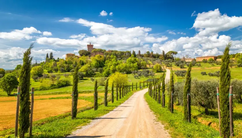 A winding dirt road lined with cypress trees leads to a Tuscan village on a hill under a blue sky.