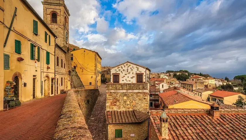 Un vicolo con case gialle e torre campanaria a Montescudaio. Cosa vedere: un borgo medievale con tetti rossi e cielo nuvoloso.