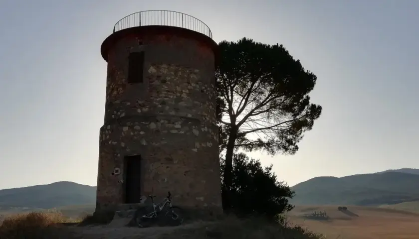 Torre circolare in pietra con una bicicletta appoggiata, vicina a un pino solitario. Paesaggio collinare sullo sfondo, forse la macchia della Magona.