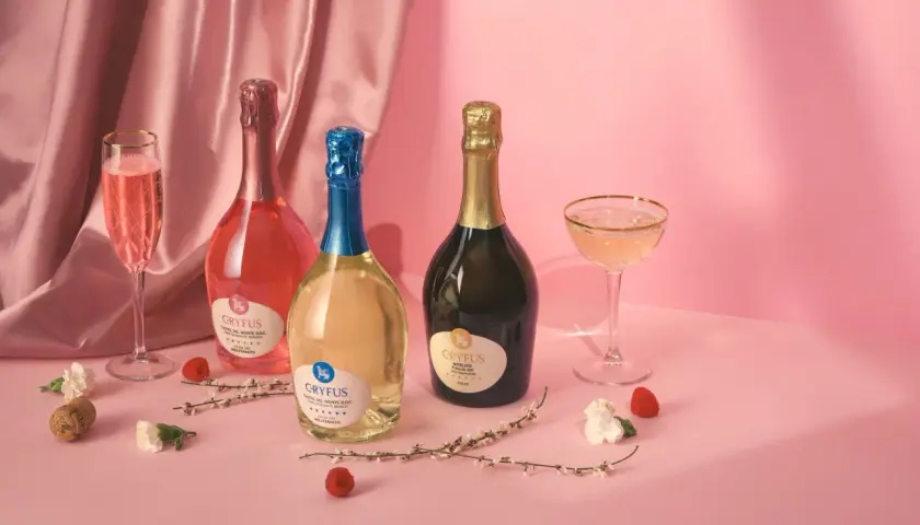 Tre bottiglie di champagne italiano, una rosata, una bianca e una dorata, con calici e decorazioni floreali su sfondo rosa.