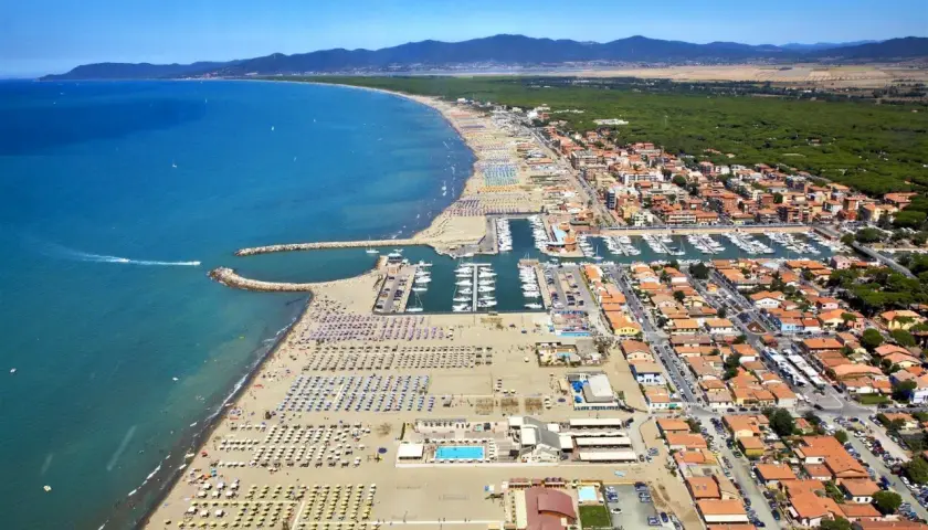 Vista aerea di Marina di Grosseto con spiaggia affollata, porto turistico pieno di barche e case con tetti rossi.