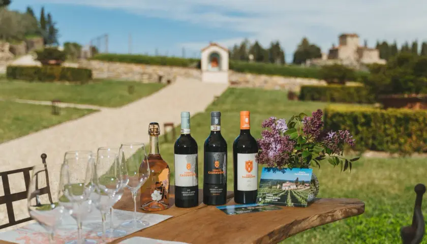 Vino e bicchieri sul tavolo di legno, con la campagna toscana e la Tenuta Casenuove sullo sfondo.