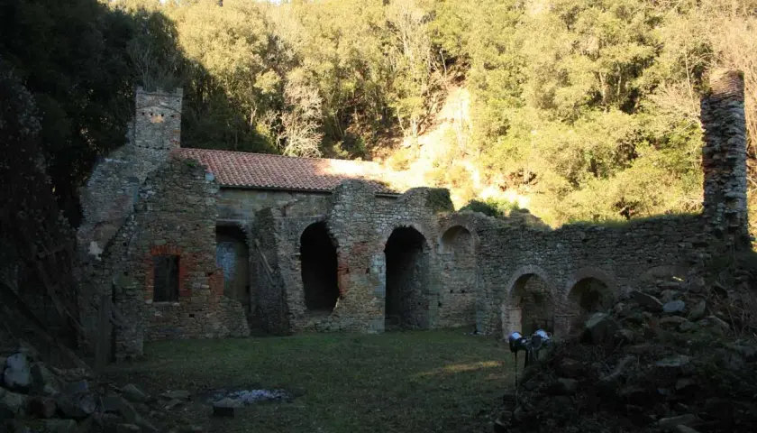 Rovine dell'eremo di San Guglielmo, con archi in pietra e un tetto parzialmente intatto, immerso nel verde di una collina boscosa.