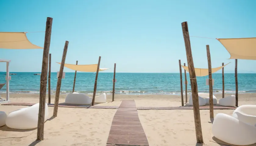 Un percorso in legno conduce al **follonica mare**, dove lettini bianchi e tende color crema invitano al relax sotto il sole.