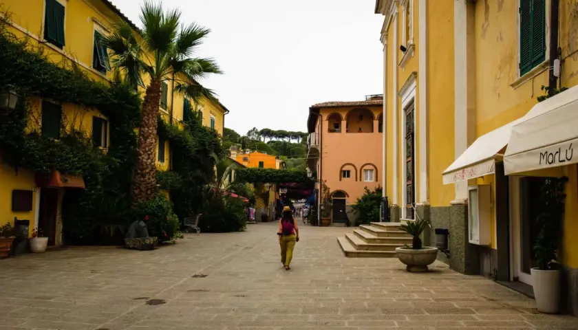 Una donna cammina in una piazza acciottolata del centro storico dell'Isola d'Elba, circondata da edifici color pastello e vegetazione lussureggiante.