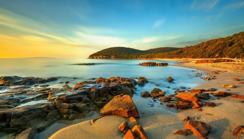 La Cala Martina al tramonto, con rocce scure e sabbia dorata bagnata da un mare calmo e cielo sfumato.