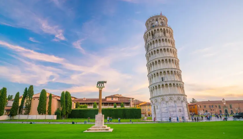 La celebre torre di Pisa, con la sua inconfondibile pendenza, si staglia contro un cielo azzurro e rosa. Un prato verde e un monumento completano la scena.